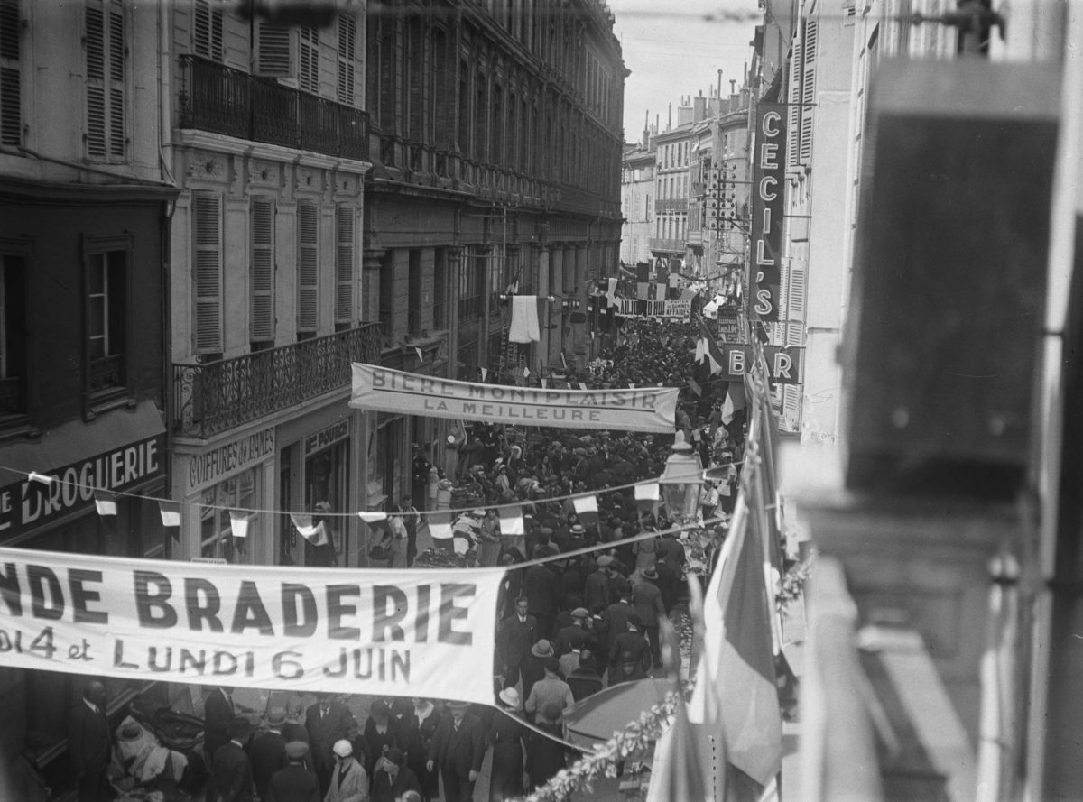 Ville de Toulouse - Archives municipales. 85Fi360 [Grande Braderie toulousaine]. Rue des Tourneurs. 4-6 juin 1932. Vue perspective plongeante - prise en direction de la rue Baronie - de la rue des Tourneurs où se presse une foule de visiteurs à l'occasion de la grande braderie toulousaine ; banderoles publicitaires dont une pour la "Bière Montplaisir - La meilleure" ; guirlandes et drapeaux ornant la rue.